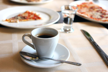 A small cup of espresso coffee, pizza and a glass of water in the background.