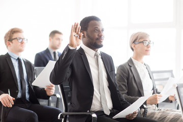Adult African-American man raising hand with question while sitting with colleagues on business meeting