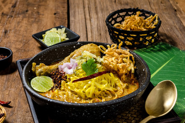 The tasty egg yellow noodle in chicken curry soup with ingredients in the bowl on wooden table. Kao Soi Kai, this is name of the traditional food of northern Thailand..