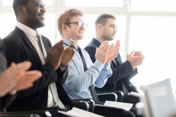Row of diverse content coworking men sitting on chairs in hall and giving applaud to speaker on business meeting