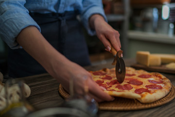 Woman cutting fresh baked homemade pizza on rustic kitchen background. Cut into slices delicious pizza with mushrooms and ham. Cheese and tomatoes on wooden table. Healthy foods, cooking concept.