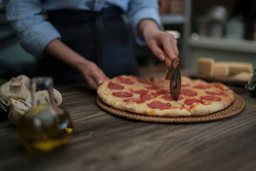 Woman cutting fresh baked homemade pizza on rustic kitchen background. Cut into slices delicious pizza with mushrooms and ham. Cheese and tomatoes on wooden table. Healthy foods, cooking concept.