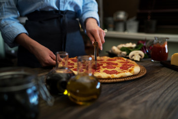 Obraz na płótnie Canvas Woman cutting fresh baked homemade pizza on rustic kitchen background. Cut into slices delicious pizza with mushrooms and ham. Cheese and tomatoes on wooden table. Healthy foods, cooking concept.