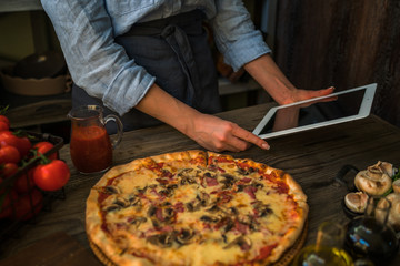 Cooking, food, technology and people concept. Young woman using a tablet computer to cook pizza in her kitchen. A woman searching the cooking pecipe and preparing food ingredient before cooking.