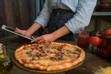 Cooking, food, technology and people concept. Young woman using a tablet computer to cook pizza in her kitchen. A woman searching the cooking pecipe and preparing food ingredient before cooking.