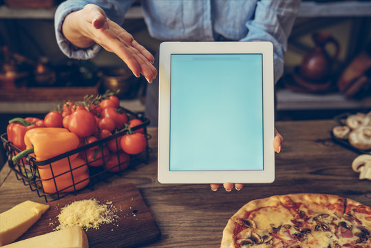 Hand Holding A Tablet Computer With White Screen. Woman Hands Showing Empty Screen Of Modern Digital Tablet. Cooking And Digital Everyday Life Online Concept. Screen In Focus With Woman In Kitchen.