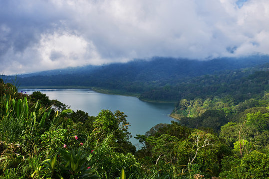 View of a lake surrounded by volcanic mountains, tropical landscape with dramatic clouds in the sky.  Lake and mountain view from a hill. Forest on the shore of a lake. Nature background.