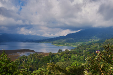 Fototapeta na wymiar Forested mountain slope in low lying cloud with the evergreen tropical trees, shrouded in mist in a scenic landscape. Low cloud shrouds the distant mountains above the waters of the lake.