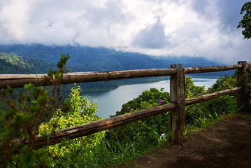 Fototapeta na wymiar Wooden fence along the shore of the lake. Low cloud shrouds the distant mountains. Tropical landscape with dramatic clouds in the sky. Lake and mountain view from a hill. Nature background.