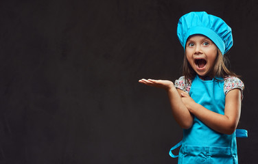 Surprised little girl dressed in blue cook posing in a studio. Isolated on dark textured background.
