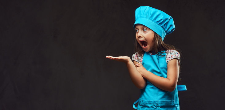 Surprised little girl dressed in blue cook posing in a studio. Isolated on dark textured background.