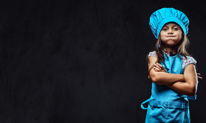 Disgruntled little girl dressed in blue cook uniform standing with crossed arms. Isolated on dark...