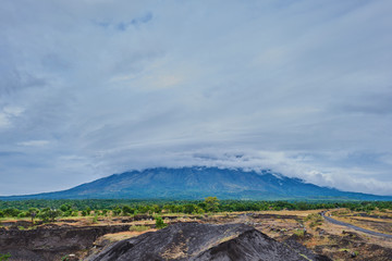 Picturesque view at the volcano peak covered in clouds. Remains of lava and a quarry with a road are at the foot of the mountain.