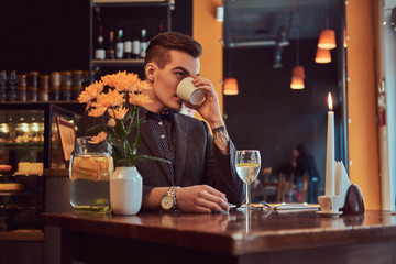 Portrait of a handsome man with stylish beard and hair dressed in a black suit sitting at a cafe, drinks morning coffee.