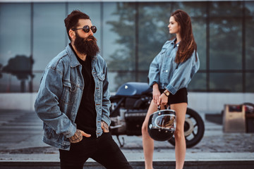Attractive hipster couple - bearded brutal male in sunglasses dressed in a jeans jacket and his young sensual girl standing on steps against a skyscraper.