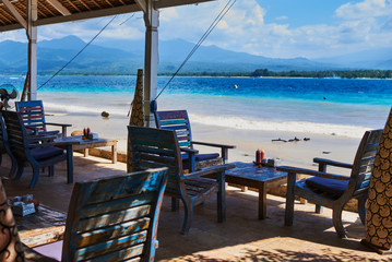 Leisure, travel and tourism concept - close up of table setting at empty open-air restaurant on the paradise beach. Wooden armchairs  on the background of a shining turquoise ocean. Tropical summer.