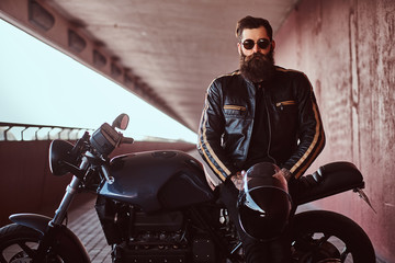 Fototapeta na wymiar Stylish fashionable biker dressed in a black leather jacket with sunglasses holds a helmet sitting on his custom-made retro motorcycle, looking at camera.