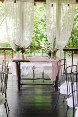 Wedding. Banquet. The festive table for guests, decorated with a composition of white and pink flowers and greenery, there are candles, served with crockery in wooden summer house.