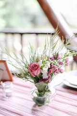 Wedding. Banquet. The festive table for guests, decorated with a composition of white and pink flowers and greenery, there are candles, served with crockery in wooden summer house.