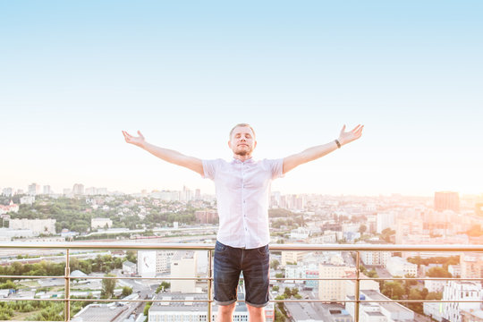 Man's portrait on a high rise building balcony overlooking city with hands rised up to sky, feeling and celebrating freedom, victory, sucsess. Expressing his joy of life. Positive emotion. Copy space.