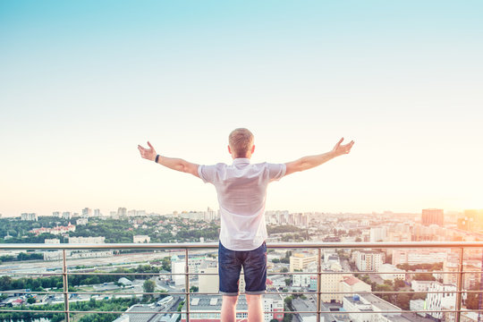 Back view Man on a high rise building balcony overlooking city with hands rised up to sky, feeling and celebrating freedom, victory, sucsess. Expressing his joy of life. Positive emotions. Copy space.