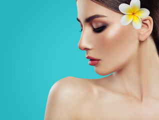 Fototapeta na wymiar Spa woman face closeup. Attractive spa model with healthy skin and tropical flower on blue background