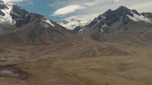 Aerial cinematic view of mountains with snow cover summit. Abra La Raya Cusco Puno Chile. Drone rising up filming mountains and valley between during cloudy day.