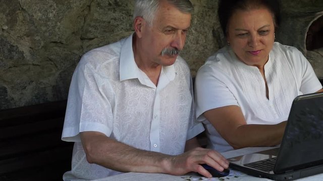 senior couple mature man and woman do web online shopping on laptop. both in white shirt staring at laptop. static medium side outdoor shot - Powered by Adobe