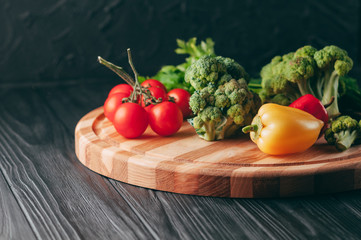 Fresh vegetables: cherry tomatoes, broccoli, pepper, dill, parsley on a round wooden board, on a dark wooden table. Recipe. Ingredients. Dietary food. Place under the text. View from above