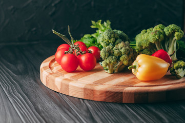 Fresh vegetables: cherry tomatoes, broccoli, pepper, dill, parsley on a round wooden board, on a dark wooden table. Recipe. Ingredients. Dietary food. Place under the text. View from above