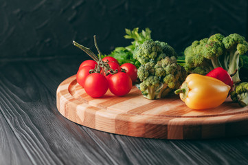Fresh vegetables: cherry tomatoes, broccoli, pepper, dill, parsley on a round wooden board, on a dark wooden table. Recipe. Ingredients. Dietary food. Place under the text. View from above