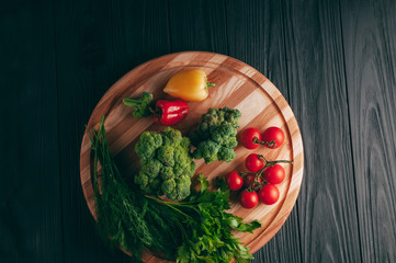 Fresh vegetables: cherry tomatoes, broccoli, pepper, dill, parsley on a round wooden board, on a dark wooden table. Recipe. Ingredients. Dietary food. Place under the text. View from above