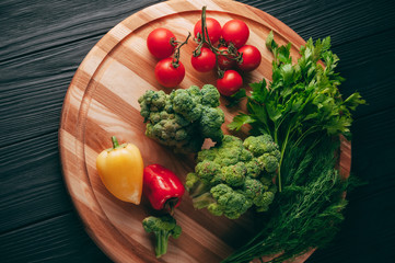 Fresh vegetables: cherry tomatoes, broccoli, pepper, dill, parsley on a round wooden board, on a dark wooden table. Recipe. Ingredients. Dietary food. Place under the text. View from above