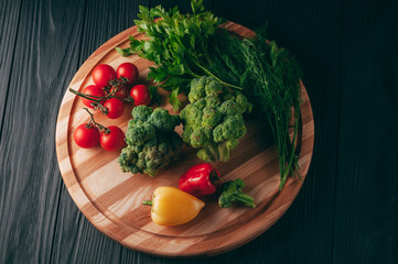 Fresh vegetables: cherry tomatoes, broccoli, pepper, dill, parsley on a round wooden board, on a dark wooden table. Recipe. Ingredients. Dietary food. Place under the text. View from above