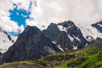 Obraz premium Picturesque landscape with snowy mountains under blue cloudy sky in summer. Green grassland in highlands near dark rocks. Majestic nature.