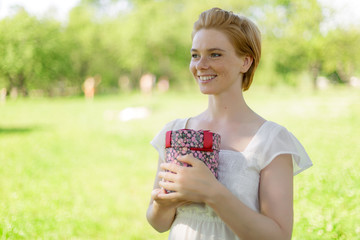 This is for you. Attractive young woman holding a gift box in her hands standing against summer green park