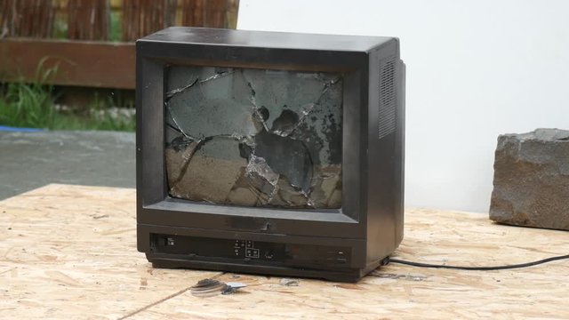 Retro black CRT TV being smashed in by a thrown rock and smoking in slow motion