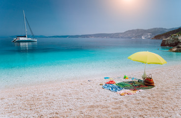 Umbrella and beach accessories on pebble beach on lazy summer day under bright sun light. Sailing boat stays at anchor in calm crystal clear blue sea water