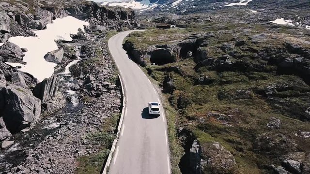 A white Nissan car is driving through the beautiful nature of Norway. This shot was taken in Stranda region which is close to Geiranger at the end of June. Snow is still visible up in the mountains.