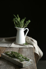 Wooden board and jug with fresh green rosemary on table. Aromatic herbs