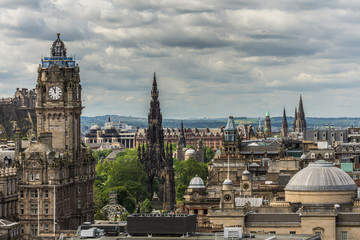 Obraz premium Edinburgh, Scotland, UK - June 13, 2012: Looking from Calton Hill upon the Balmoral Clock Tower along Princess street. Black Scott Monument. Green hills on horizon. Heavy Cloudscape.