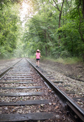 Young girl in hat walking down railroad tracks in the fog after a rain shower with rays of sunlight shining through trees