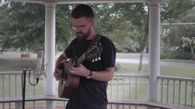 Man playing guitar and singing under a gazebo in the rain - Push in from the side shot