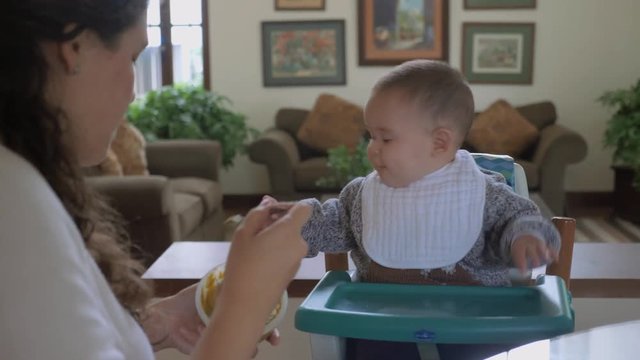 Mother feeding his baby boy with green vegetables pap in living room at home. Winter time clothing. Child routine, day-to-day activities. Pan shot.