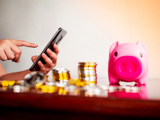 hand holding and pointing index finger to touch smart phone with rolls of many coin and piggy box.Concept of save money,investment plan,earning from internet,payment or shopping online.Selective focus