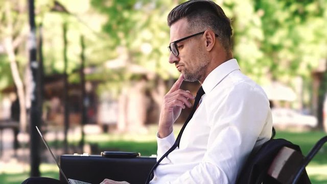 Side view of calm businessman dressed in shirt using laptop computer while sitting outside on a bench