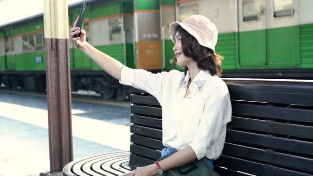 Happy Asian woman passenger with casual taking selfie by smart mobile phone in train station rails for travel in Bangkok city, Thailand. lifestyle and transportation concept.