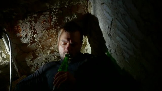 drunk alcoholic man lying in a ruined building in the dark and drinking beer from a bottle. degradation of the young man's personality