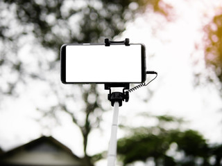 taking selfie picture by smart phone with selfie stick on blurred beautiful tree plant and forest background vacation, selective focus on phone isolated white screen smartphone concept.