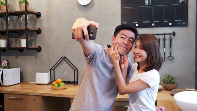 Happy young Asian couple using smartphone for selfie while cooking in the kitchen at home. Man and woman preparing healthy food. Lifestyle couple at home concept.